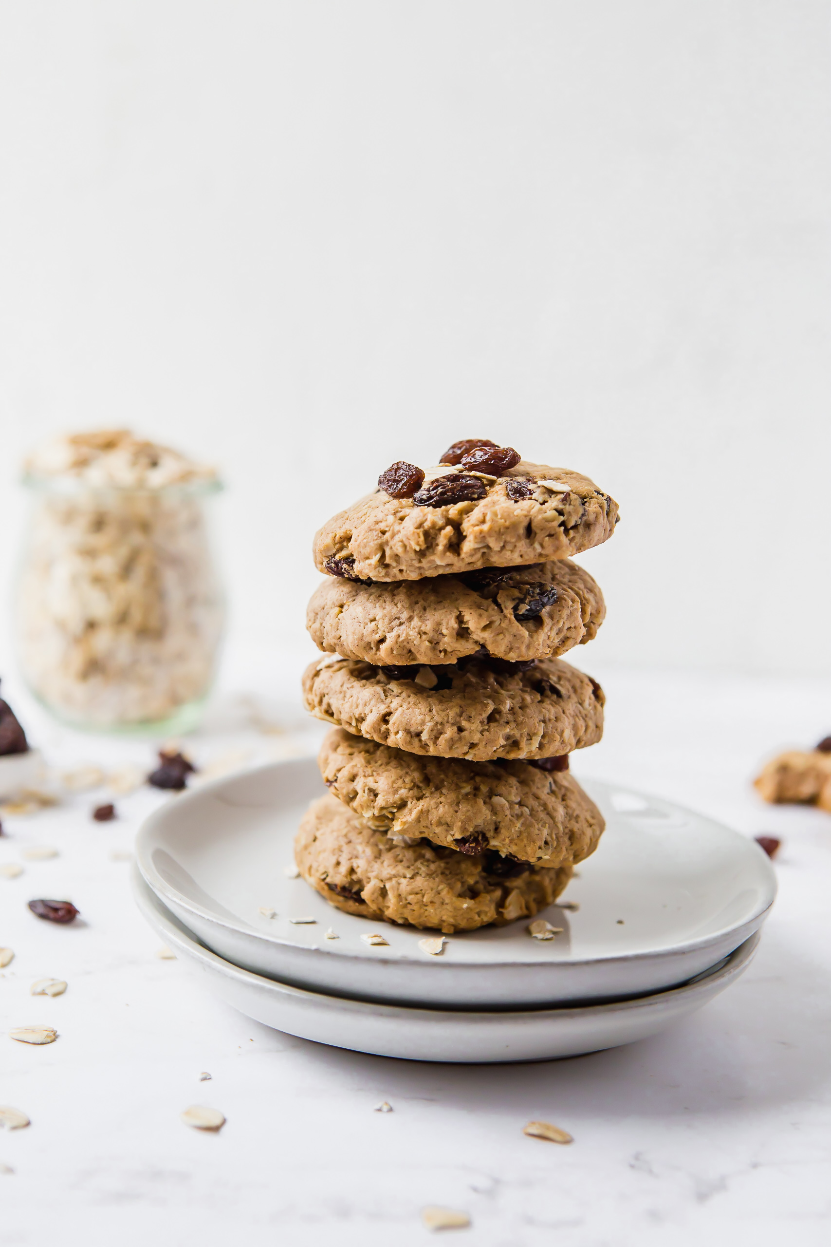 Five oatmeal raisin cookies stacked on top of each other then places on two small plates. There is a small jar of raw oats in the background on the left.