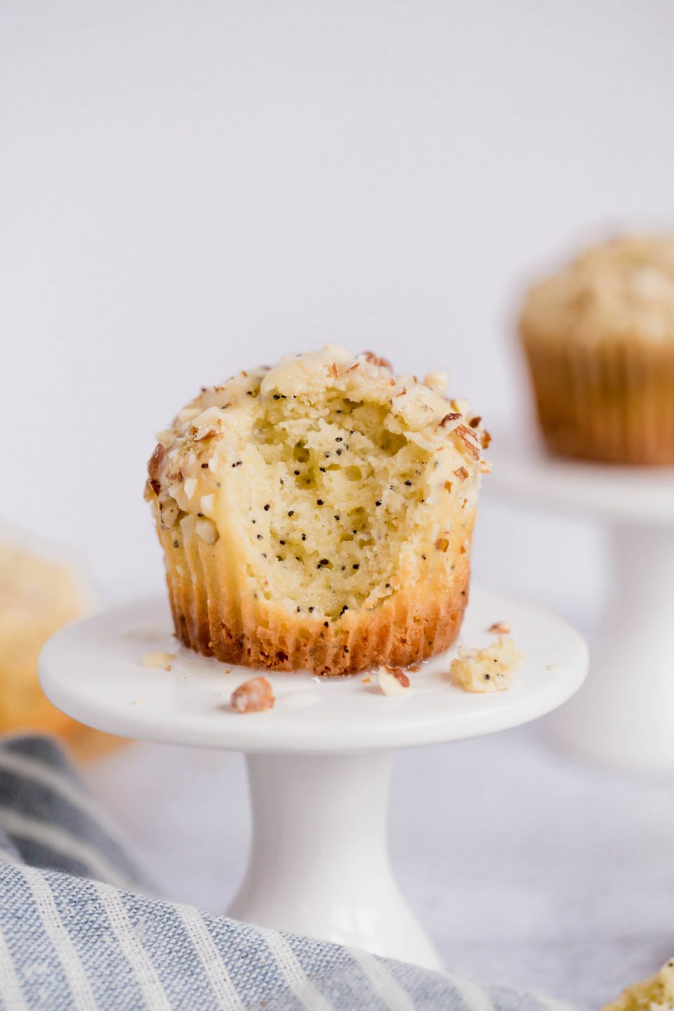 A front on shot of two gluten-free almond poppyseed muffins on small white cupcake stands. The front muffin has a large bite taken out of the top front. 