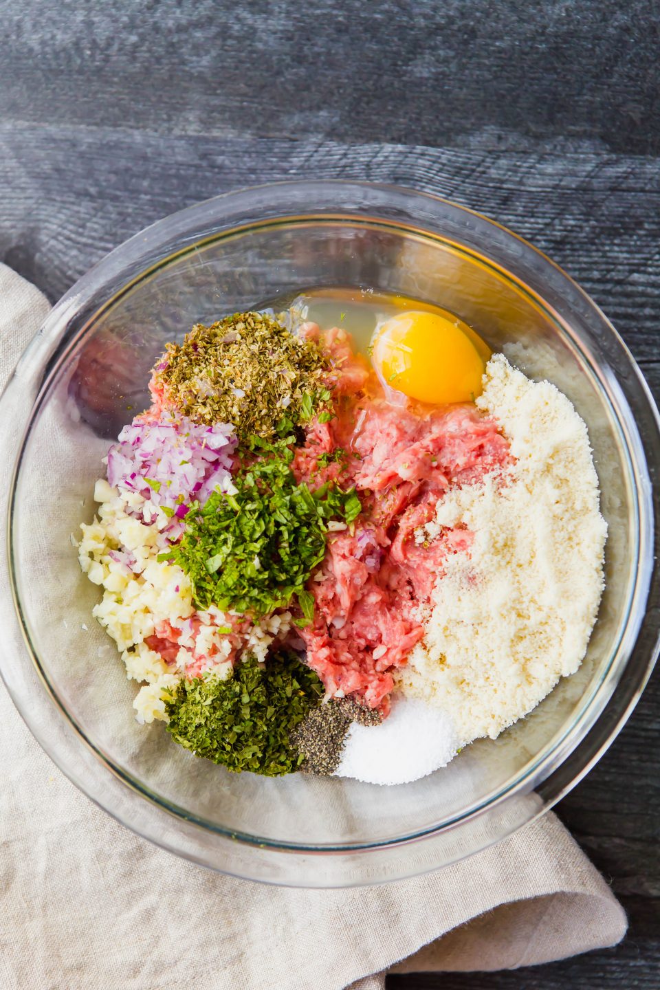 An overheard shot of a bowl with raw meatball ingredients including ground lamb, almond flour, egg, salt, pepper, red onion, oregano, garlic, and parsley. 