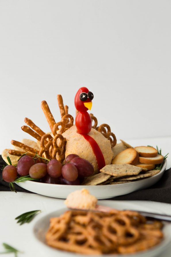 A cheeseball shaped into a ball with baby tomatoes on a stick to make it look like a turkey head. Pretzel twists and sticks are stuck in the back of the cheeseball to make it look like turkey feathers.