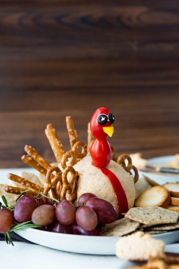 Cheeseball shaped to look like a turkey. Pretzel twists and sticks are placed in the back to look like turkey feathers. A few tomatoes on a toothpick make a turkey head. Plate shows crackers and grapes to dip in the cheese.