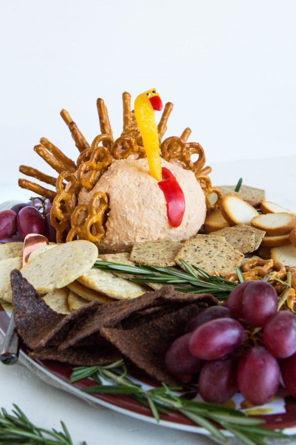 Photos of a cheeseball platter with lots of crackers and grapes with rosemary as garnish. The cheeseball is made to look like a turkey with a bell pepper head and pretzels for the feathers.