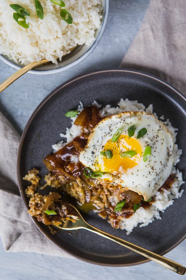 Photo of a loco loco dish on a gray plate with a bowl of rice in the background. A loco loco is white rice, hamburger patty, brown onion gravy, topped with an over-easy egg.
