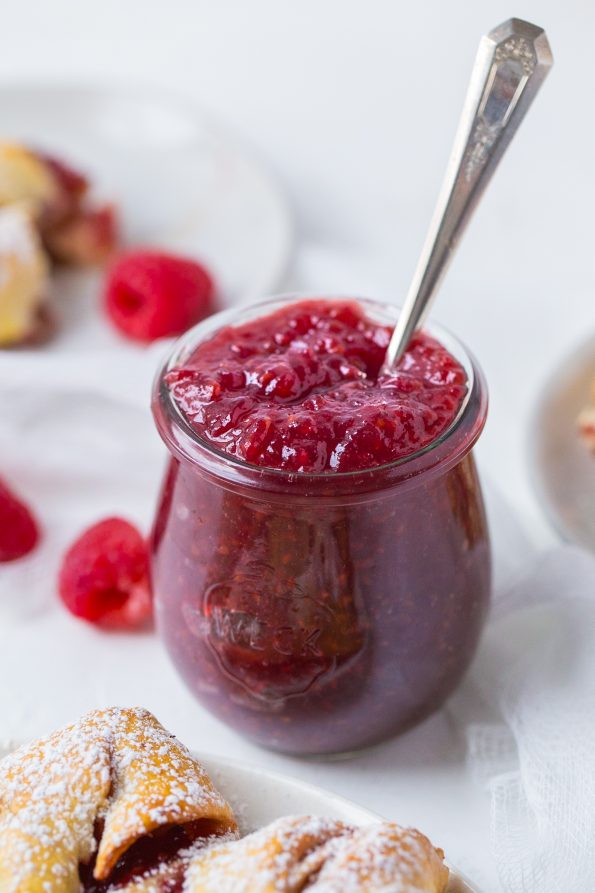 A shot of a jar of raspberry jam with a spoon sticking out of it. In the background are raspberries and pastries.