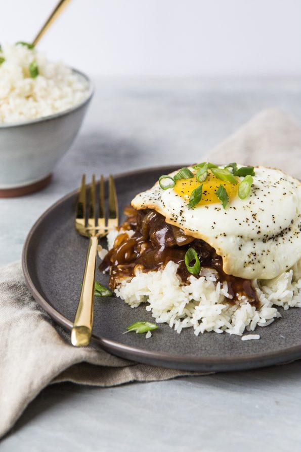 Photo of a loco loco dish on a gray plate with a bowl of rice in the background. A loco loco is white rice, hamburger patty, brown onion gravy, topped with an over-easy egg.