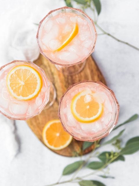Three glasses of paloma cocktails with lemon slices and ice. They are set on a wooden cutting board with sliced lemons and grapefruits scattered around.