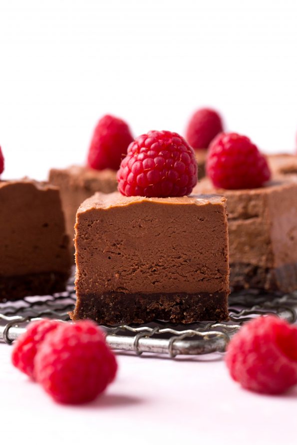 Straight on shot of chocolate cheesecake bars with a chocolate crust. Raspberries are places on top and scattered in the foreground.