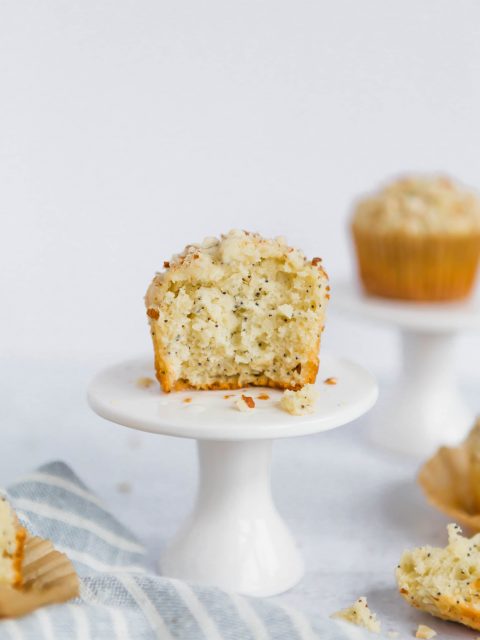 A front on shot of an almond poppyseed muffin on a small white cupcake stand. There is another cupcake in the background. In the foreground is a light-blue striped towel with pieces of muffins scattered around.