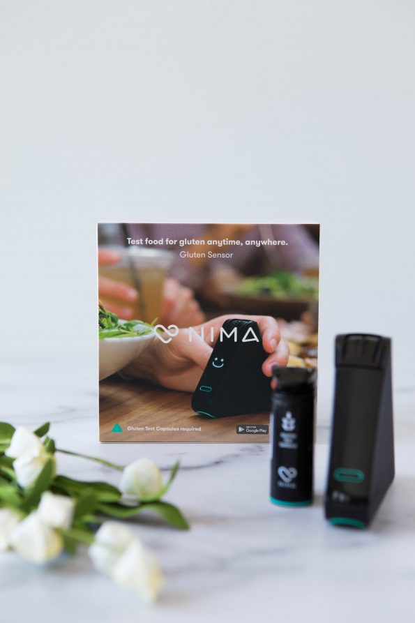 Photo of a Nima Sensor and gluten capsule with a box in the background. Flowers are placed in the front left of the photo.