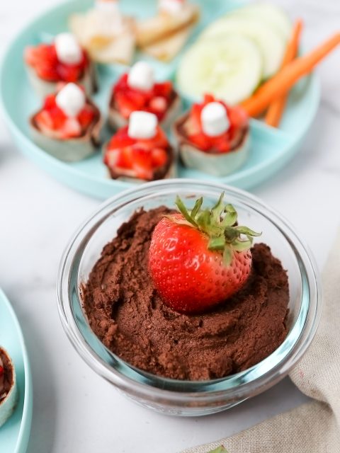 An overhead photos of homemade chocolate hummus in a small bowl with a strawberry placed in the middle. There is a kids plate with veggies and turkey slices the background.
