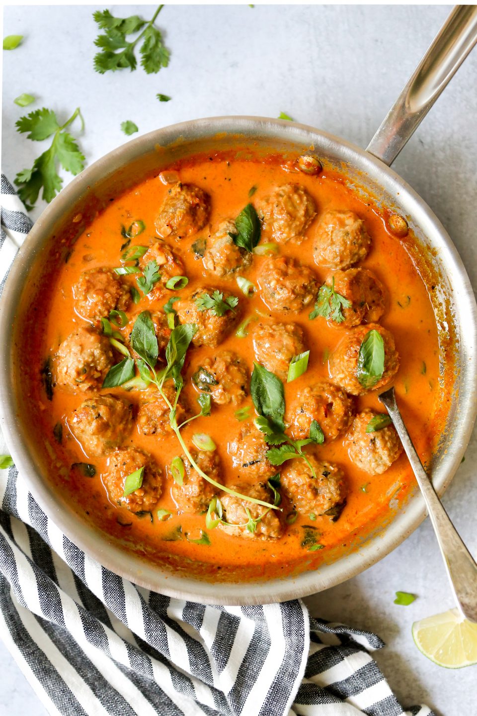 Overhead shot of turkey meatballs in a coconut curry sauce in a silver saucepan. A striped towel is draped across the bottom corner of the photo and cilantro is scattered around. 