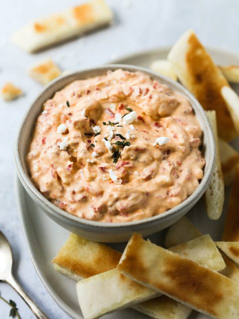 A 45 degree angle of roasted red pepper and feta dip in a gray bowl, on a gray plate with a cement background. Slices of toasted pita bread are scattered around.