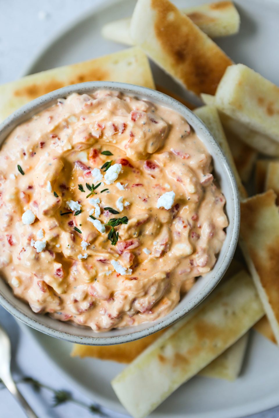 An overhead photo of roasted red pepper and feta dip in a gray bowl, on a gray plate with a cement background. Slices of toasted pita bread are scattered around.