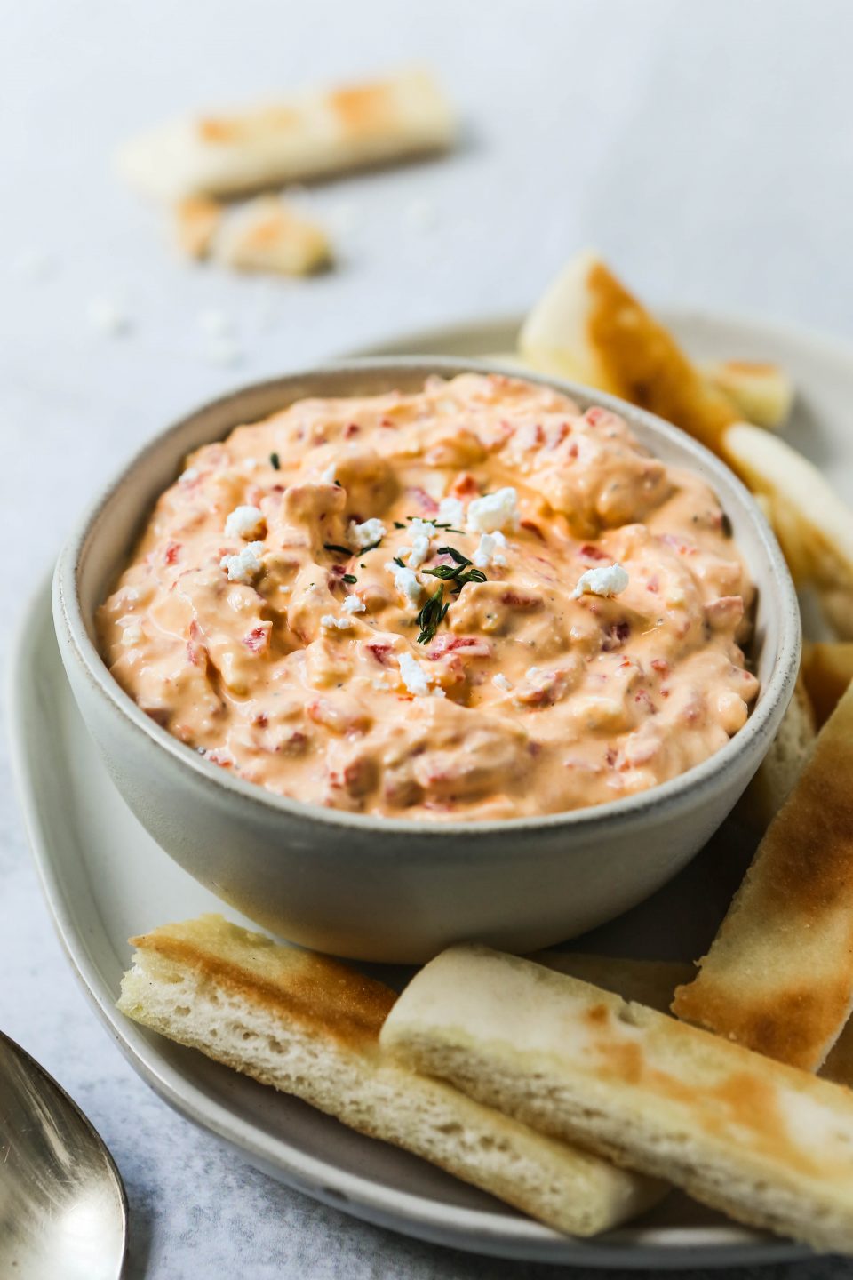 A 45 degree angle of roasted red pepper and feta dip in a gray bowl, on a gray plate with a cement background. Slices of toasted pita bread are scattered around.