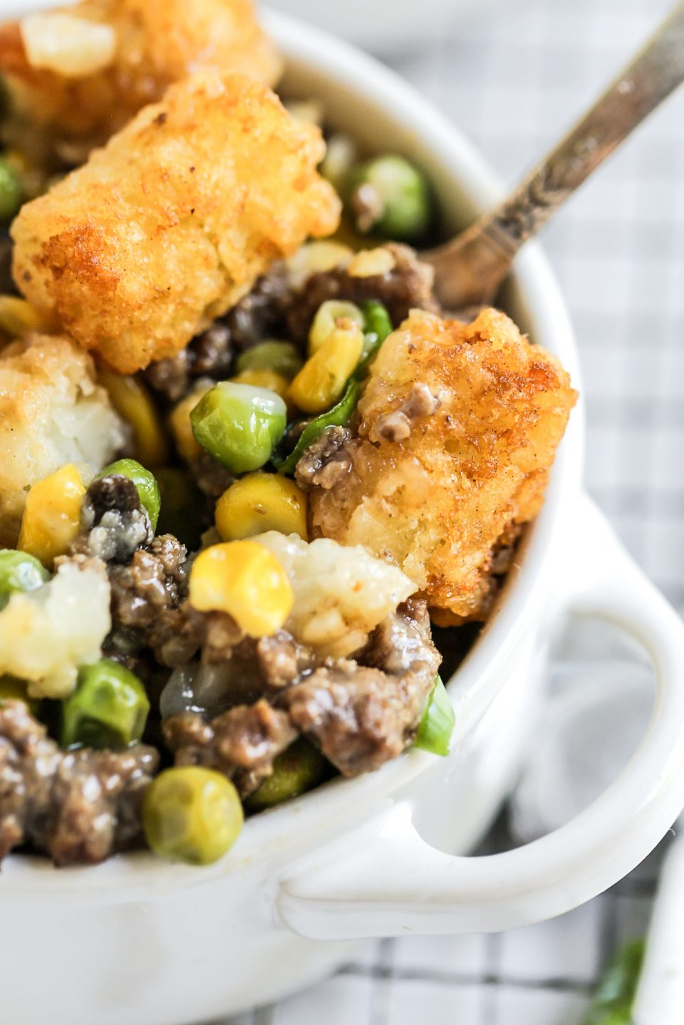 An up close photo of tator tot hot dish, including meat, tator tots, peas, and corn, in a white croquette with a fork sticking out.