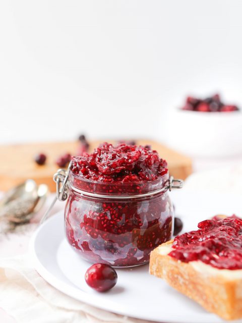 A jar overflowing with homemade cranberry jam thickened with chia seeds. There is a also a slide of toast with the jam spread on it on a plate in the foreground.