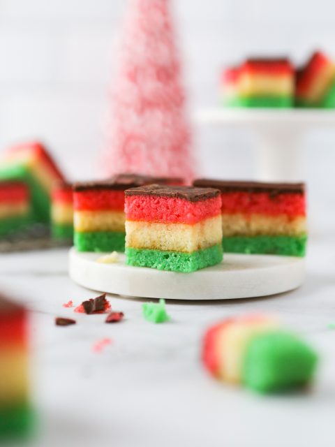 Gluten-free venetian cookies with layered red, white, and green almond sponge cake. Layered with apricot jam and coated in semi-sweet chocolate. Bite-sized cookies that are scattered on a marble surface.