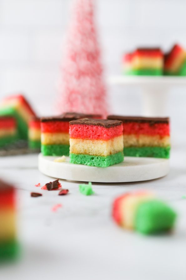 Gluten-free venetian cookies with layered red, white, and green almond sponge cake. Layered with apricot jam and coated in semi-sweet chocolate. Bite-sized cookies that are scattered on a marble surface.