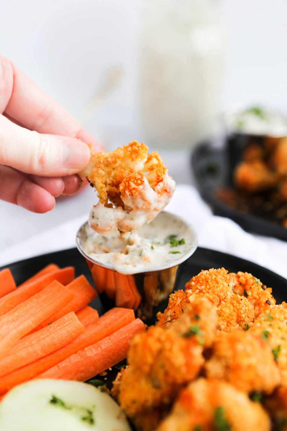 A photo of a hand holding buffalo cauliflower bites dipping into a container of dairy-free ranch dressing. 