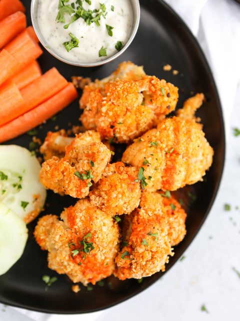 An overhead photo of buffalo cauliflower bites on a black plate. There are cucumbers, carrots, and a vegan ranch dressing in the background.