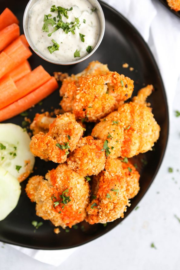 An overhead photo of buffalo cauliflower bites on a black plate. There are cucumbers, carrots, and a vegan ranch dressing in the background.
