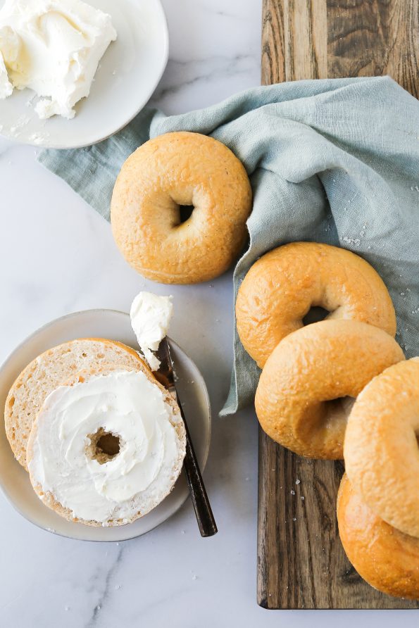 A photo of gluten-free bagels spread across a cutting board over a light blue napkin. There is one bagel cut in half smeared with cream cheese.