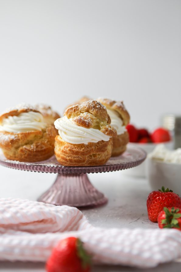Gluten-free cream puff on a light pink cake pedestal. There are strawberries scattered throughout the photo.