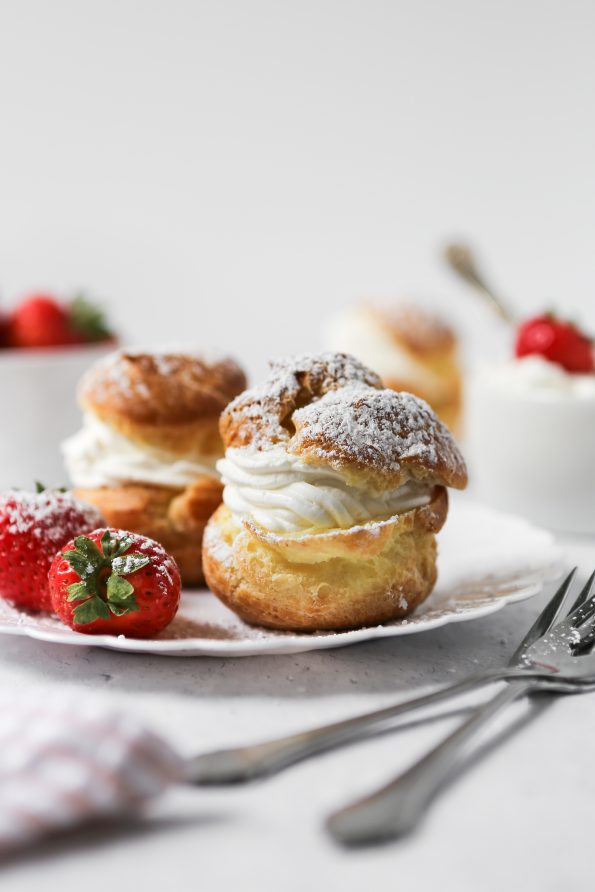 A photo of 2 gluten-free cream puffs on a plate. They are split in half and filled with fluffy whipped cream. There are strawberries placed the plate.